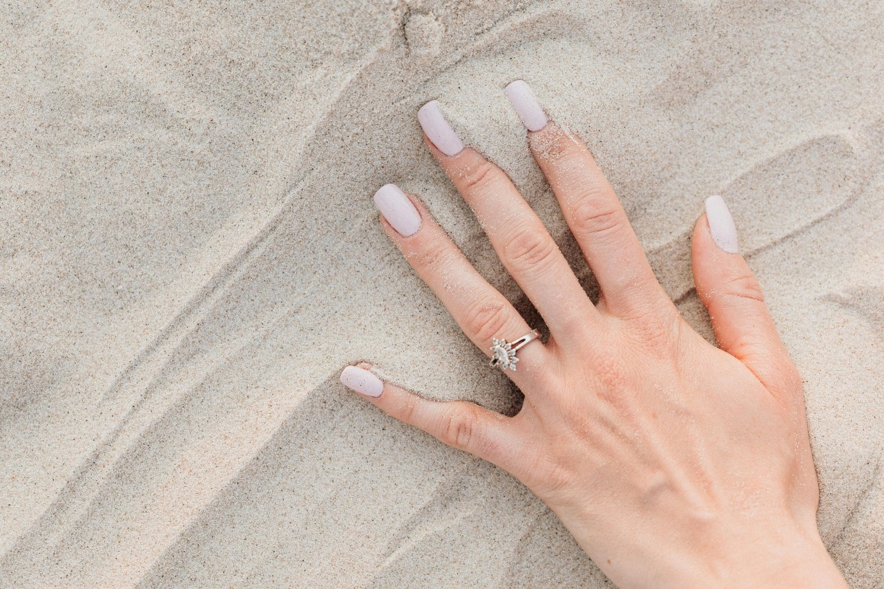 A picture of a woman’s hand wearing a distinctive engagement ring, resting on the sand.