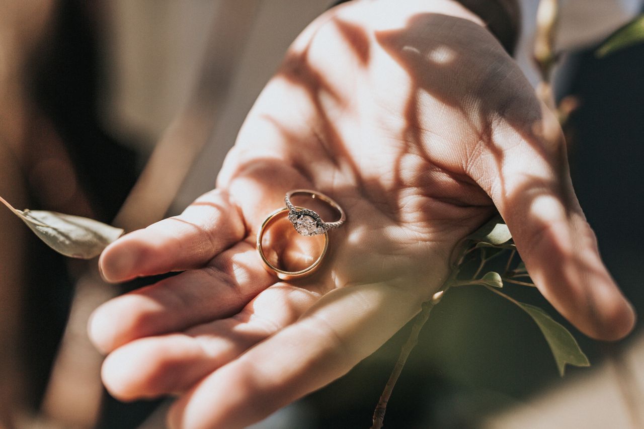 A man holding an elegant diamond engagement ring and a wedding band in the palm of his hand under dappled light.