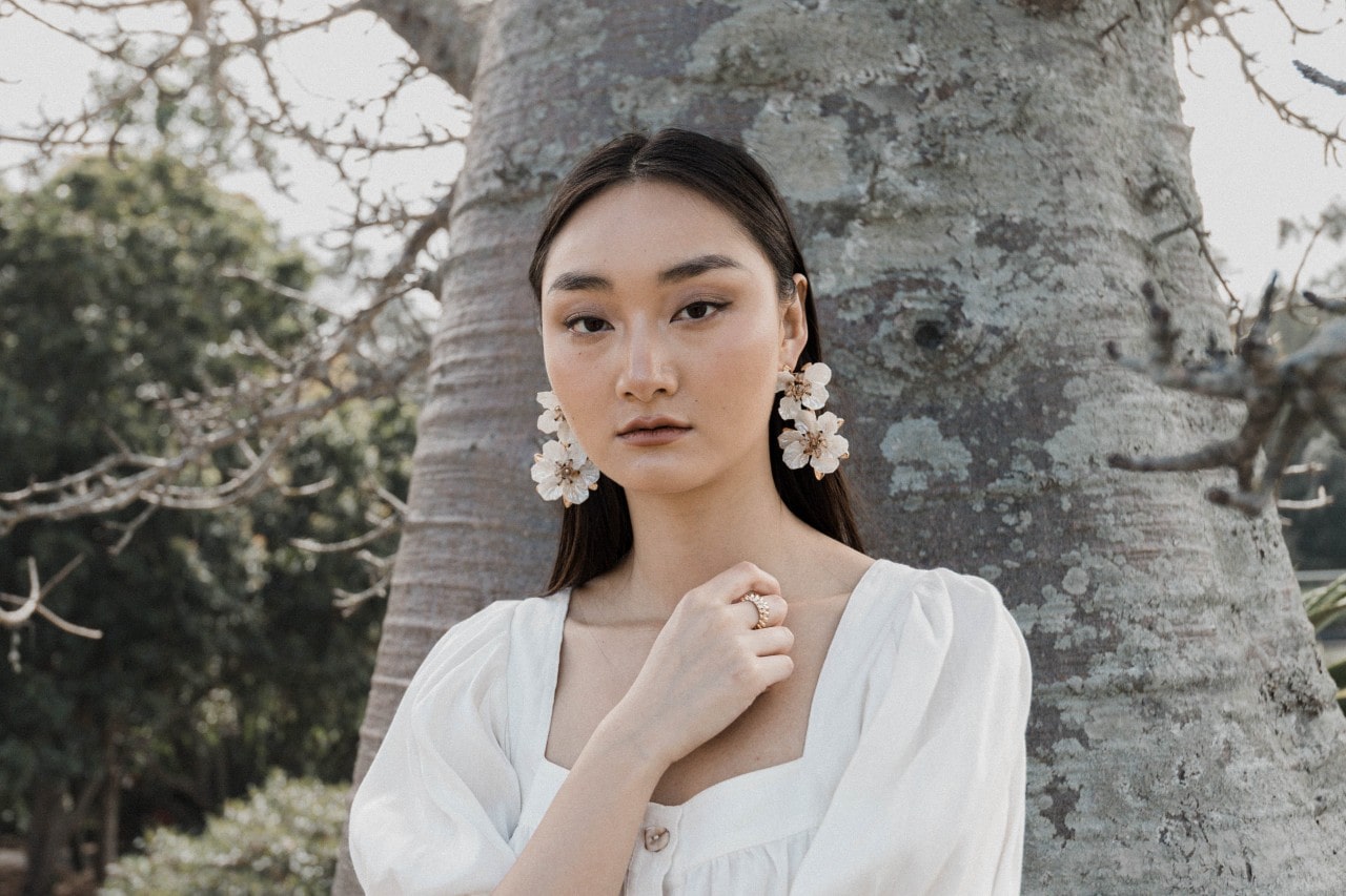 A woman wearing white floral earrings, standing in front of a tree.