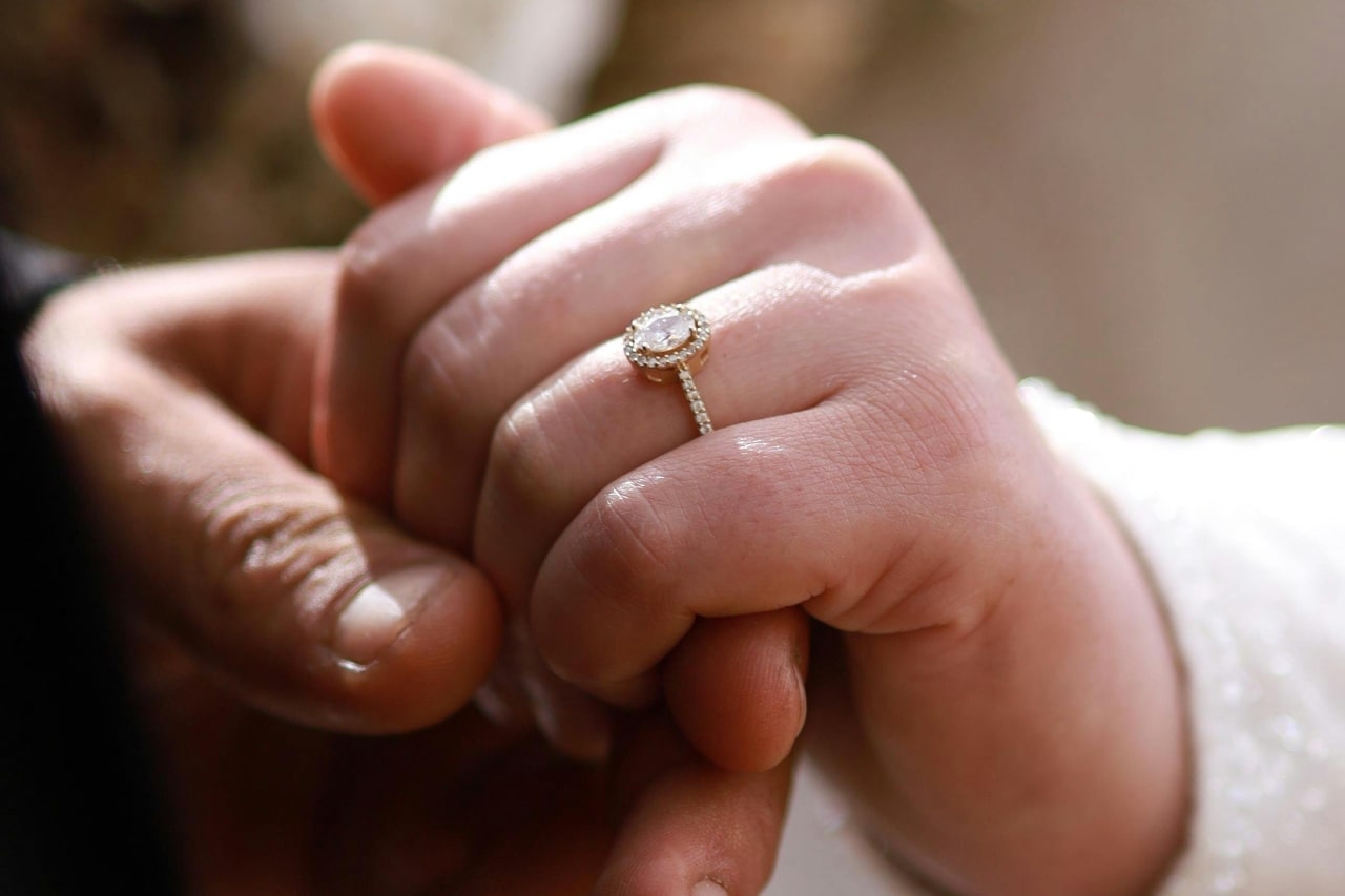 a man’s hand holding a woman’s – the woman’s adorned with an oval cut halo engagement ring