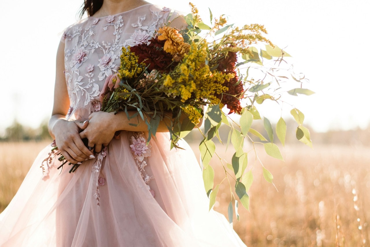 a bride standing in a field in the fall, holding a fall-themed bouquet of flowers