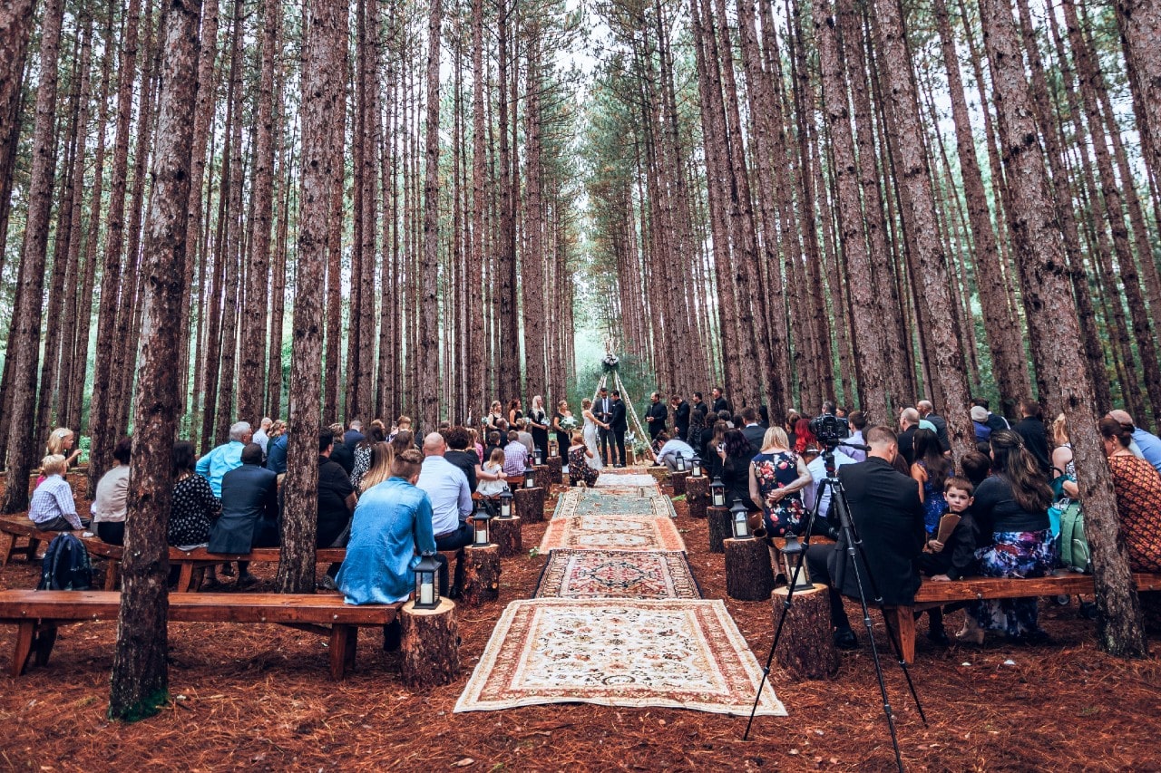 a wedding in a forest with tall trees, wooden benches, and warm-toned rugs lining the aisle