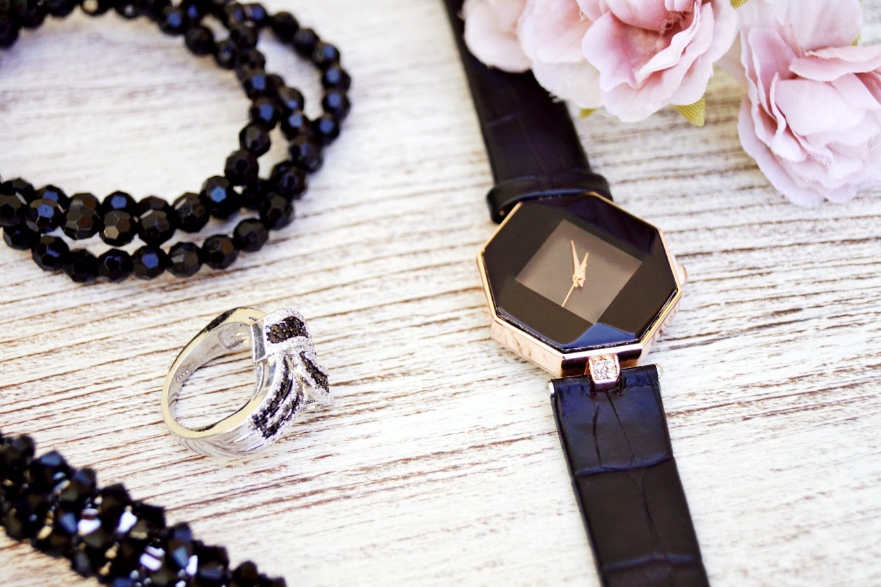 A distinctive minimalist watch displayed next to a fashionable black diamond ring and beaded necklace.