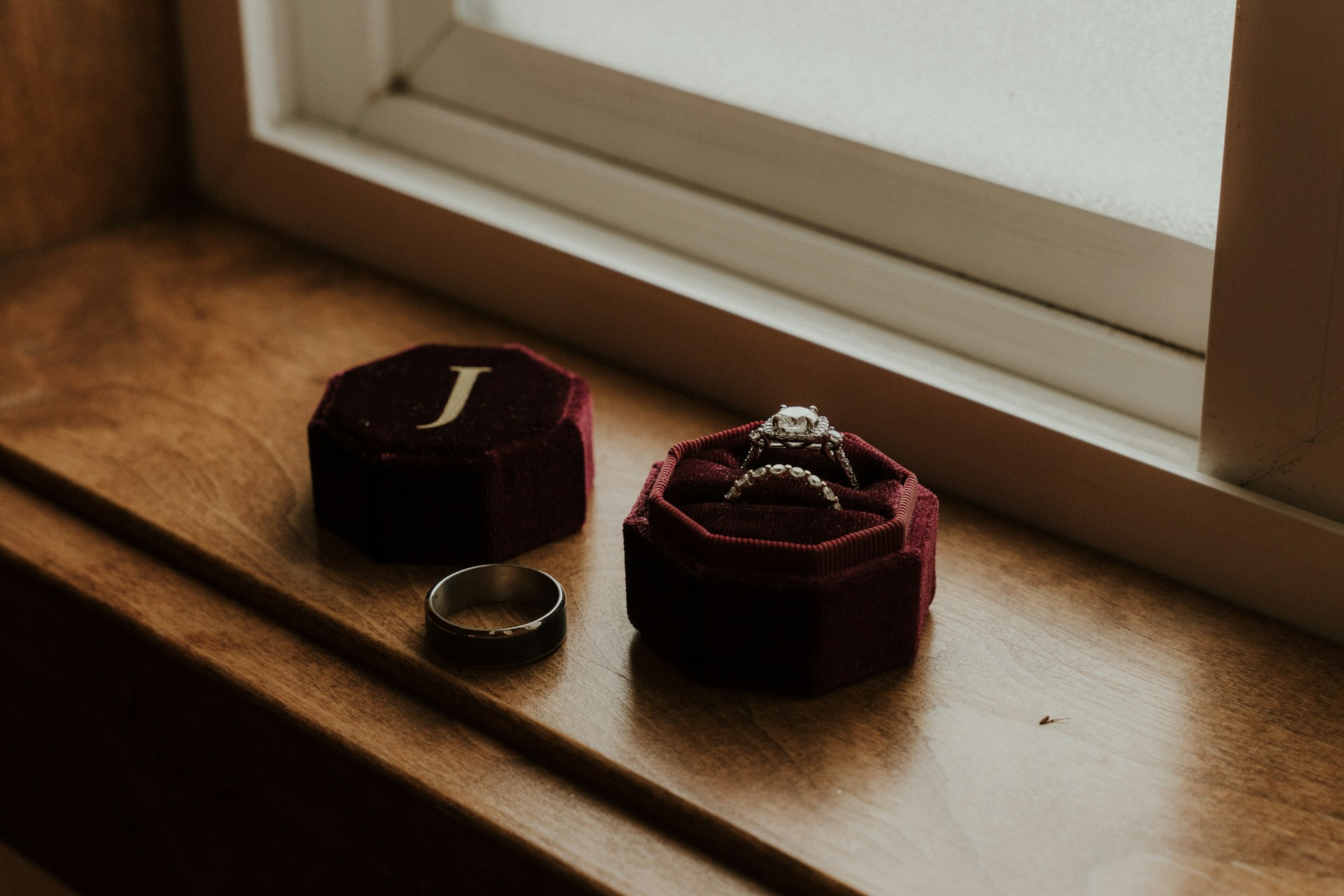 A ring box containing a bridal engagement ring and wedding band, next to a man’s wedding band on a windowsill.