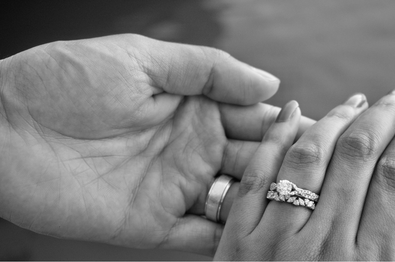 A close-up of a man’s hand holding a woman’s– his adorned with a wedding band, hers with a bridal stack.