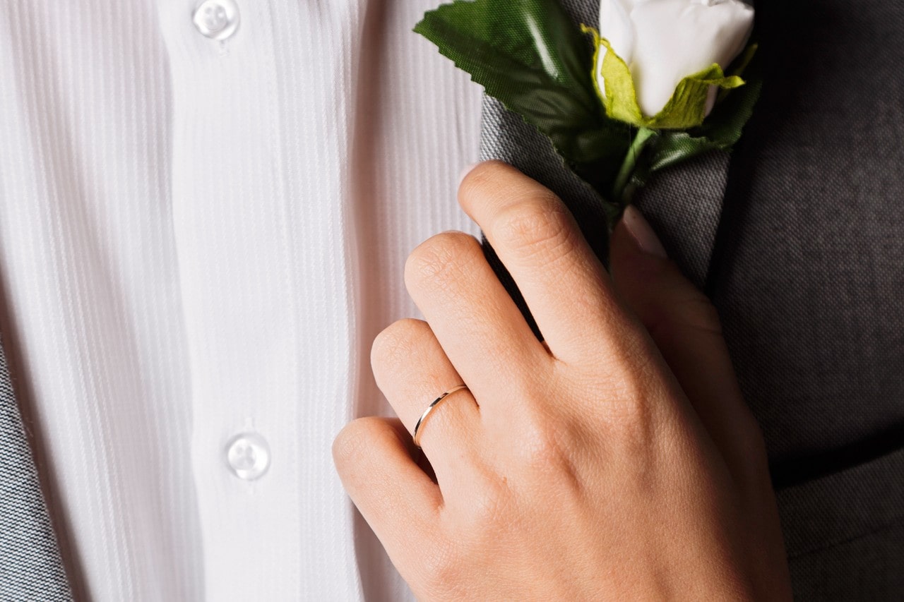 A close-up of a bride-to-be’s hand placing a boutonniere on her groom’s coat, with emphasis on the simple gold band on her finger.