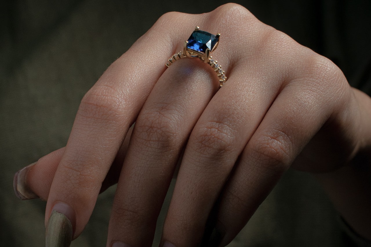 A close-up of a woman’s manicured hand, adorned with a stunning side stone engagement ring with an emerald center stone.