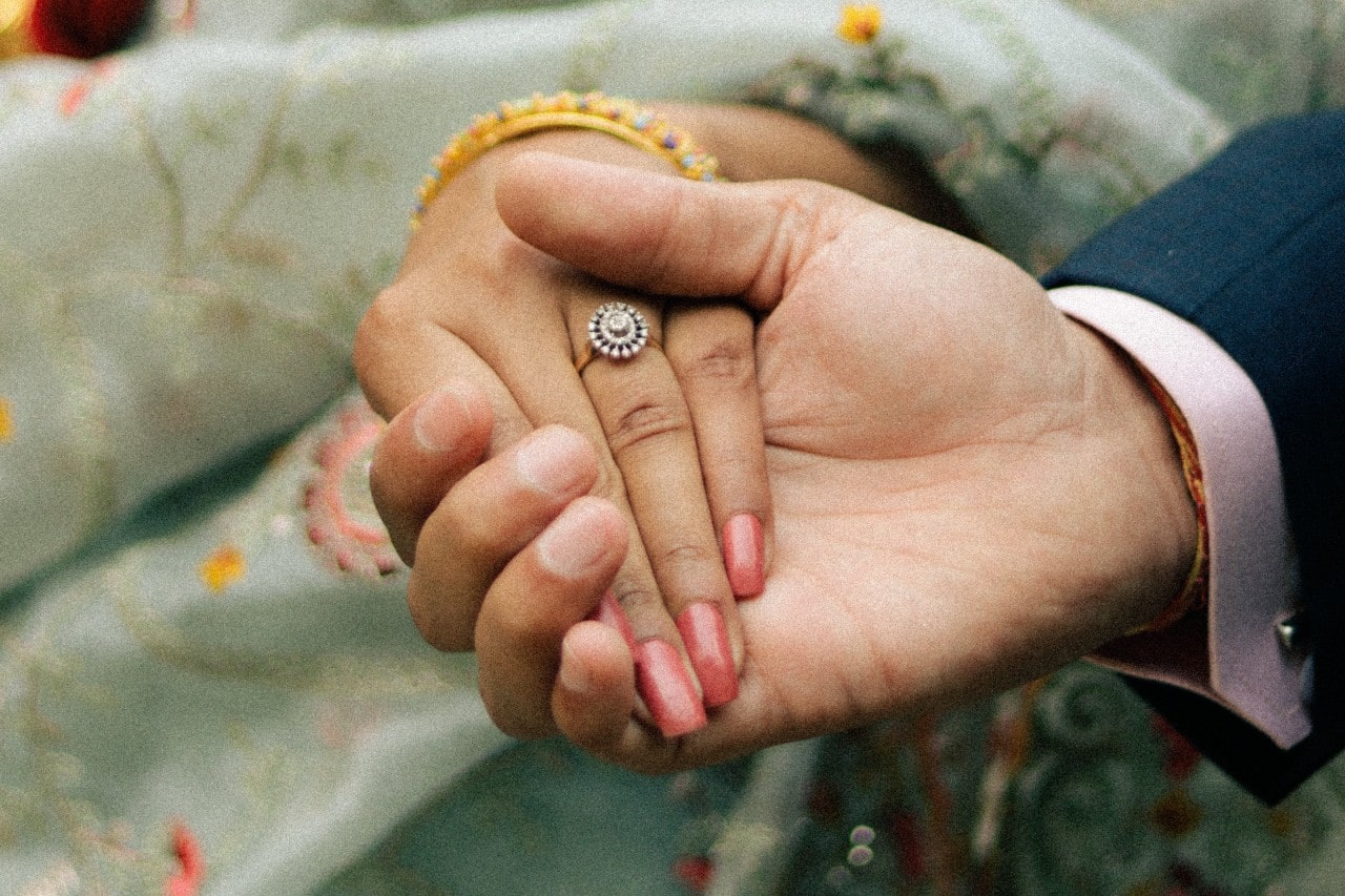 A man holding his bride-to-be’s hand wearing an ornate engagement ring.